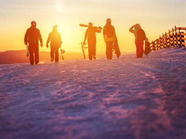 As the sun dips below the horizon, a group of adventurous friends trudges through the snow, their silhouettes framed by the golden glow of twilight. With skis and snowboards in tow, they are ready to conquer the slopes under the evening sky, sharing laughter and camaraderie on this winter escapade.